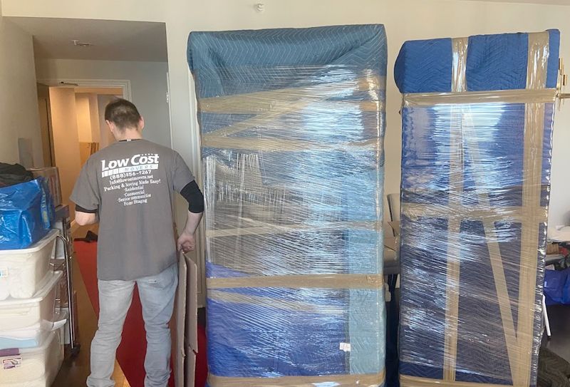 A mover wraps large furniture with blue padding and tape. Boxes and hallway are visible.