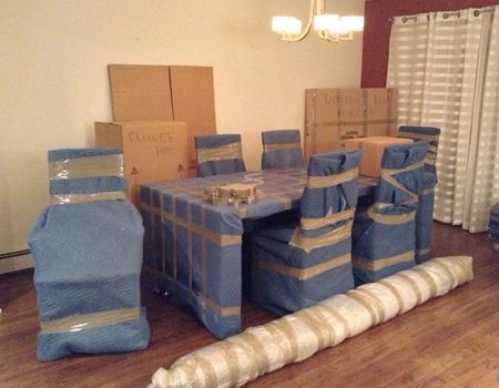 Dining table and chairs wrapped in blue padding, prepared for moving, with boxes in a room.