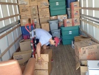 A person bends into a packed moving truck, stacking boxes. Brown cardboard boxes and green storage bins are overflowing.