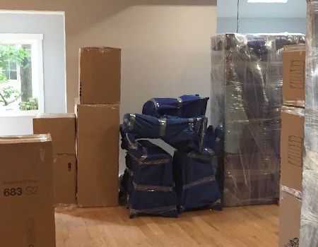 Boxes and furniture wrapped in plastic and ready for a move, inside a room with a window.