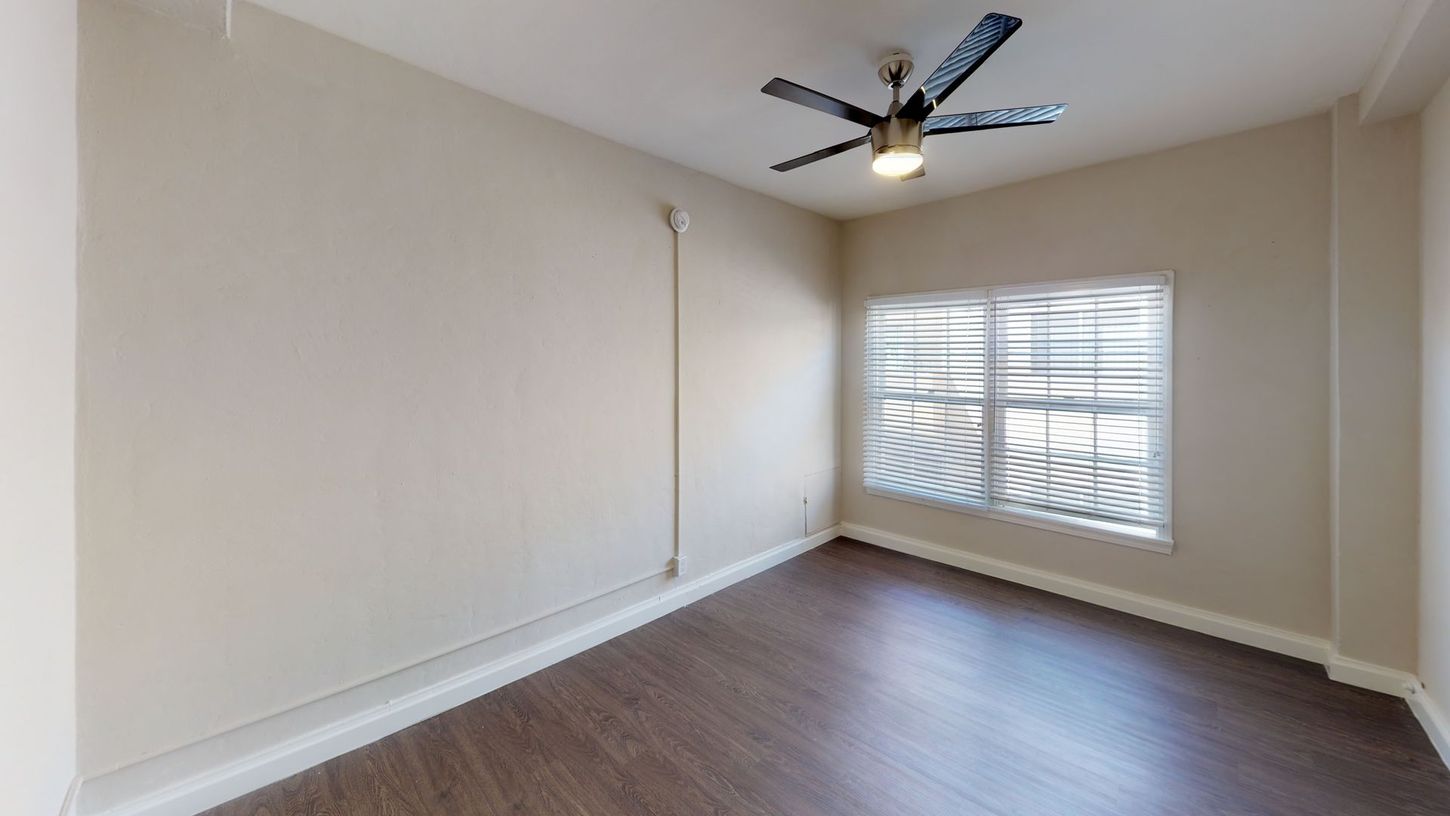 Empty bedroom with wood flooring, beige walls, and window with blinds. Ceiling fan with light.