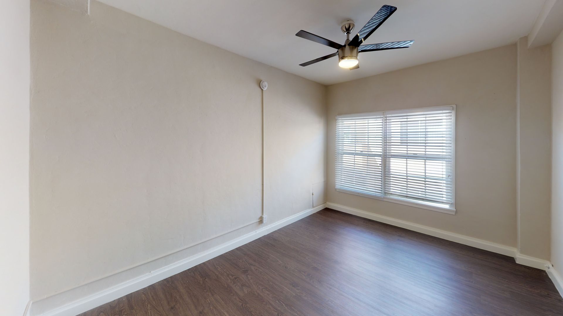 Empty bedroom with wood flooring, beige walls, and window with blinds. Ceiling fan with light.