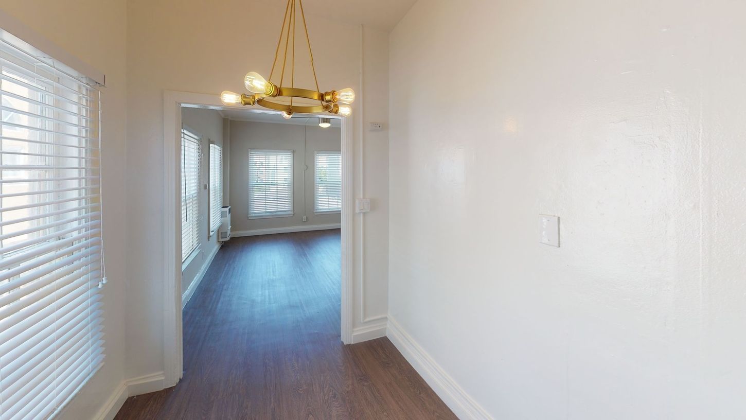 Hallway with dark wood floor leading to a room with windows; gold chandelier hangs from ceiling.