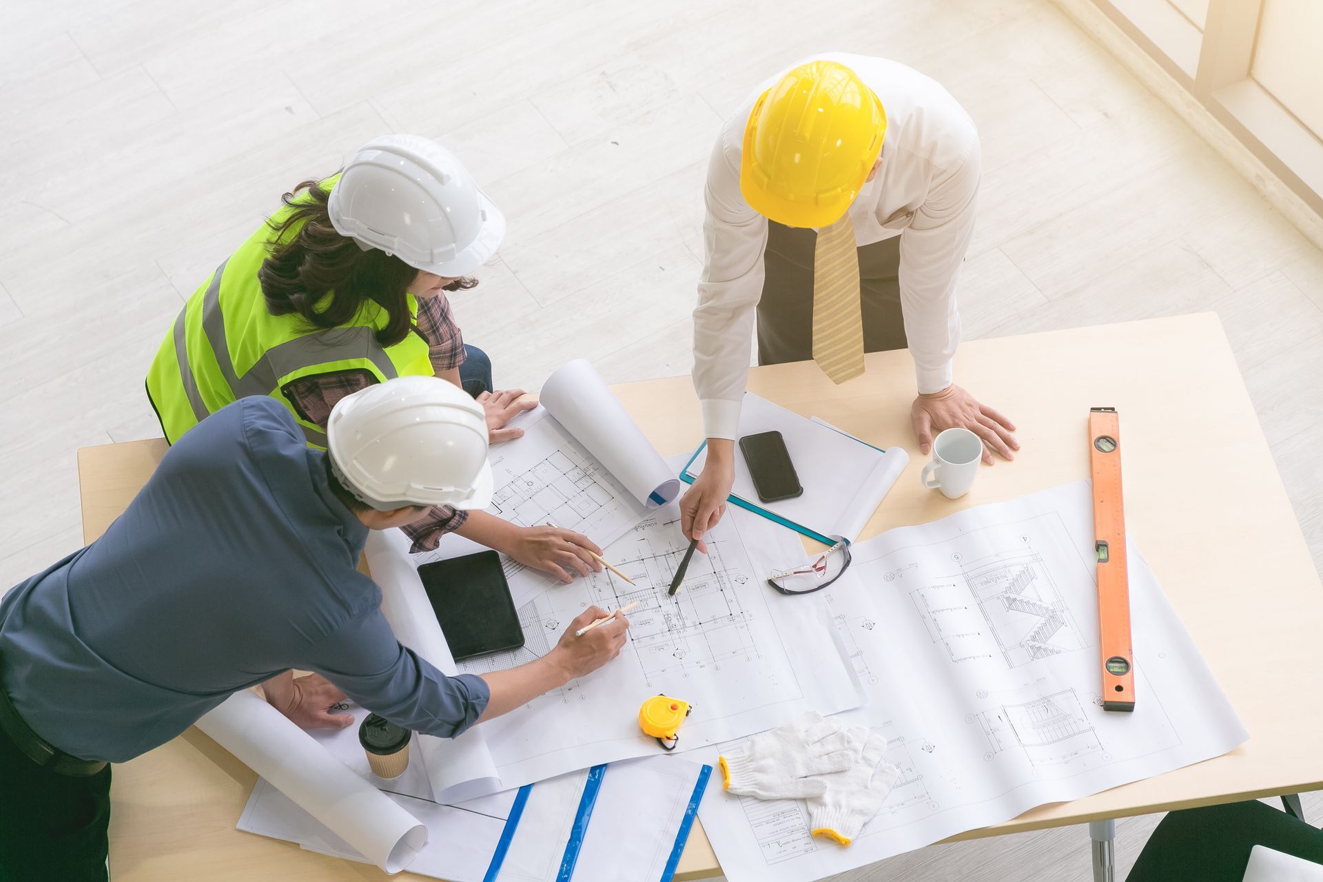 Architects in hard hats review blueprints on a desk, discussing plans, with tools and devices nearby.