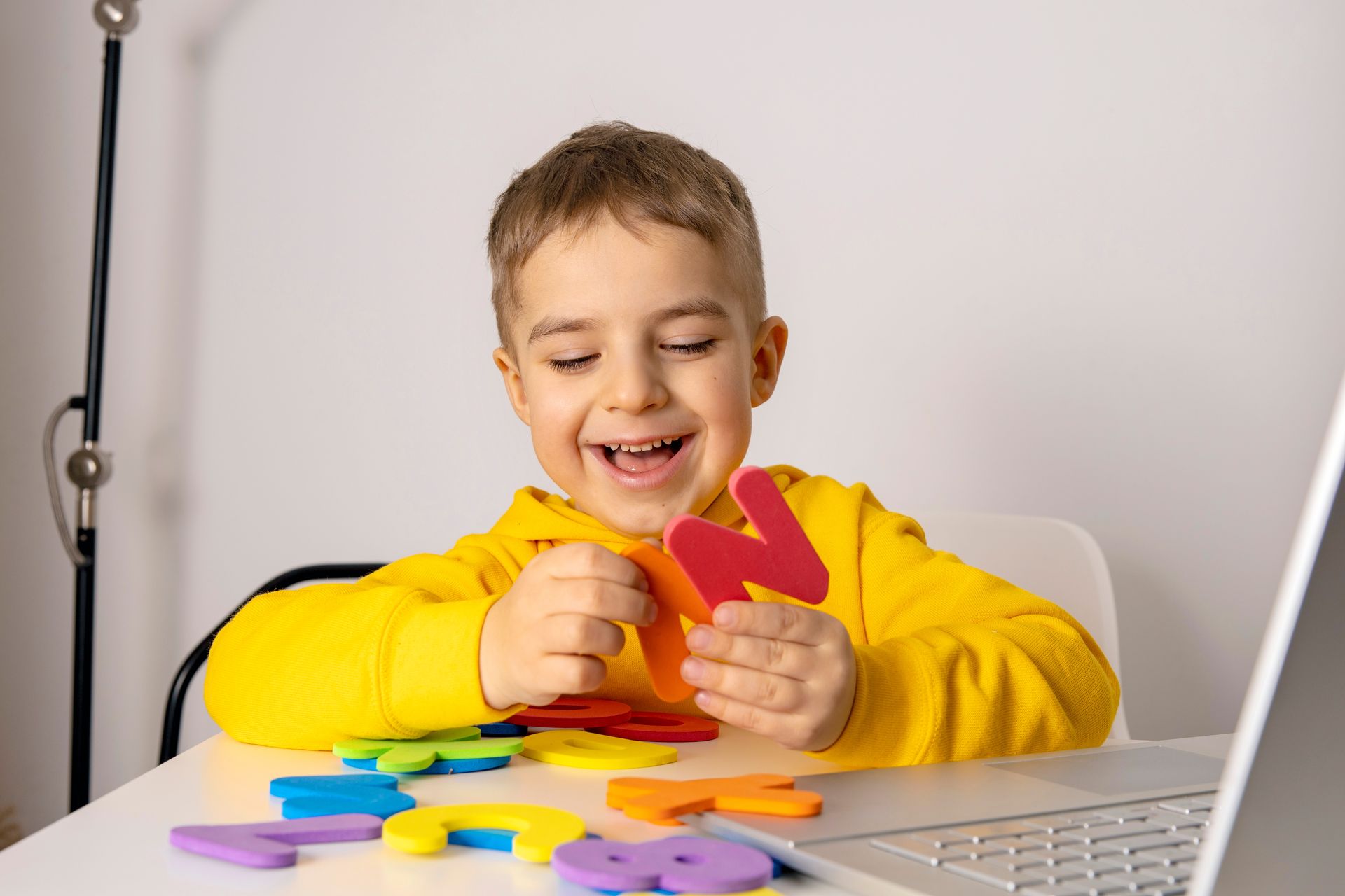 Boy in yellow hoodie playing with colorful foam letters at a desk, smiling. Laptop in frame.