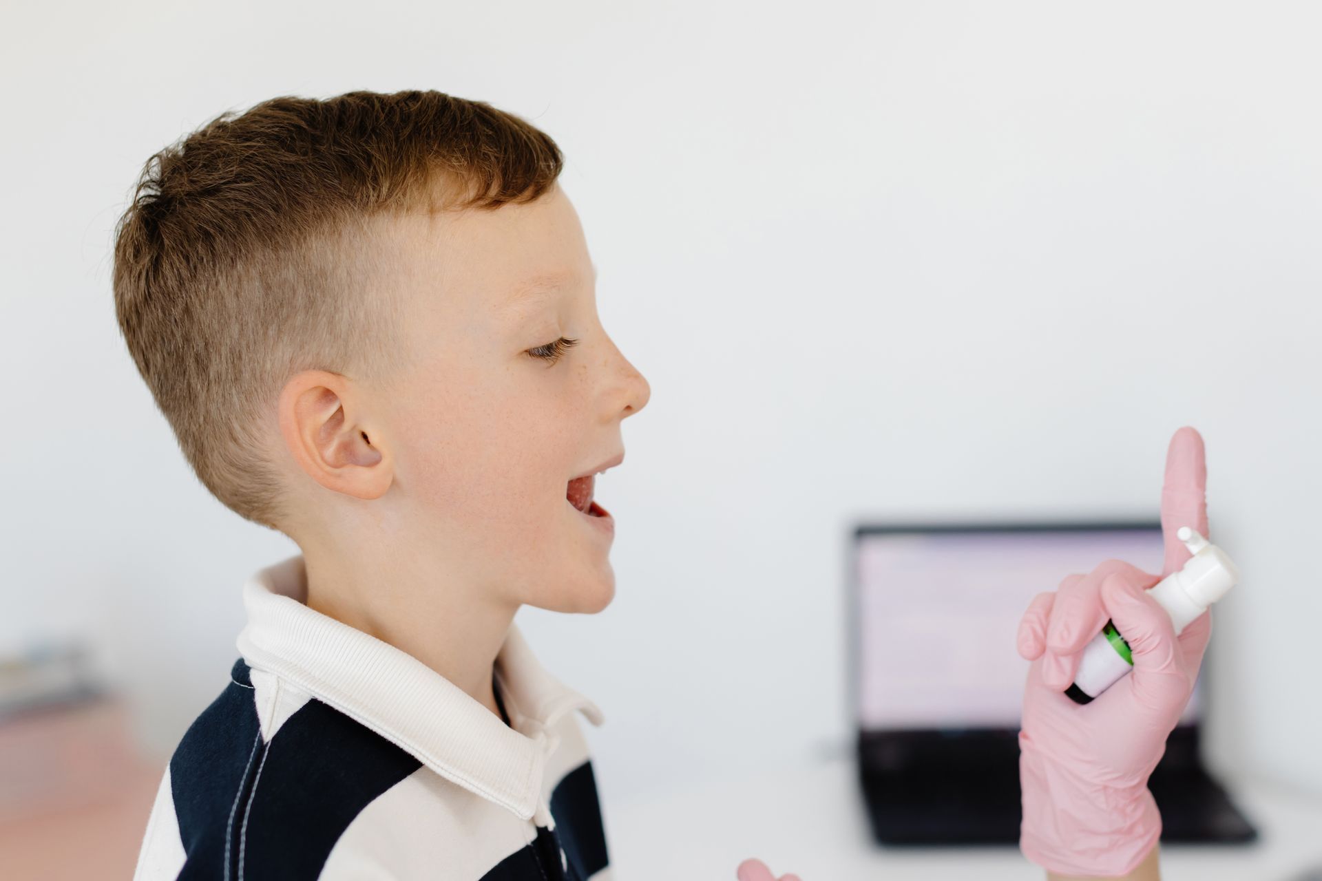 Boy receiving nasal spray, mouth open, hand with pink glove holding bottle. White background, laptop.