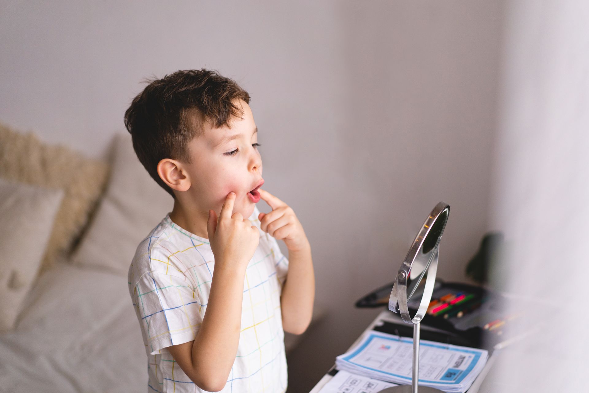 Boy in a white shirt looking in a mirror, touching his mouth. Bright room, small mirror on a table.