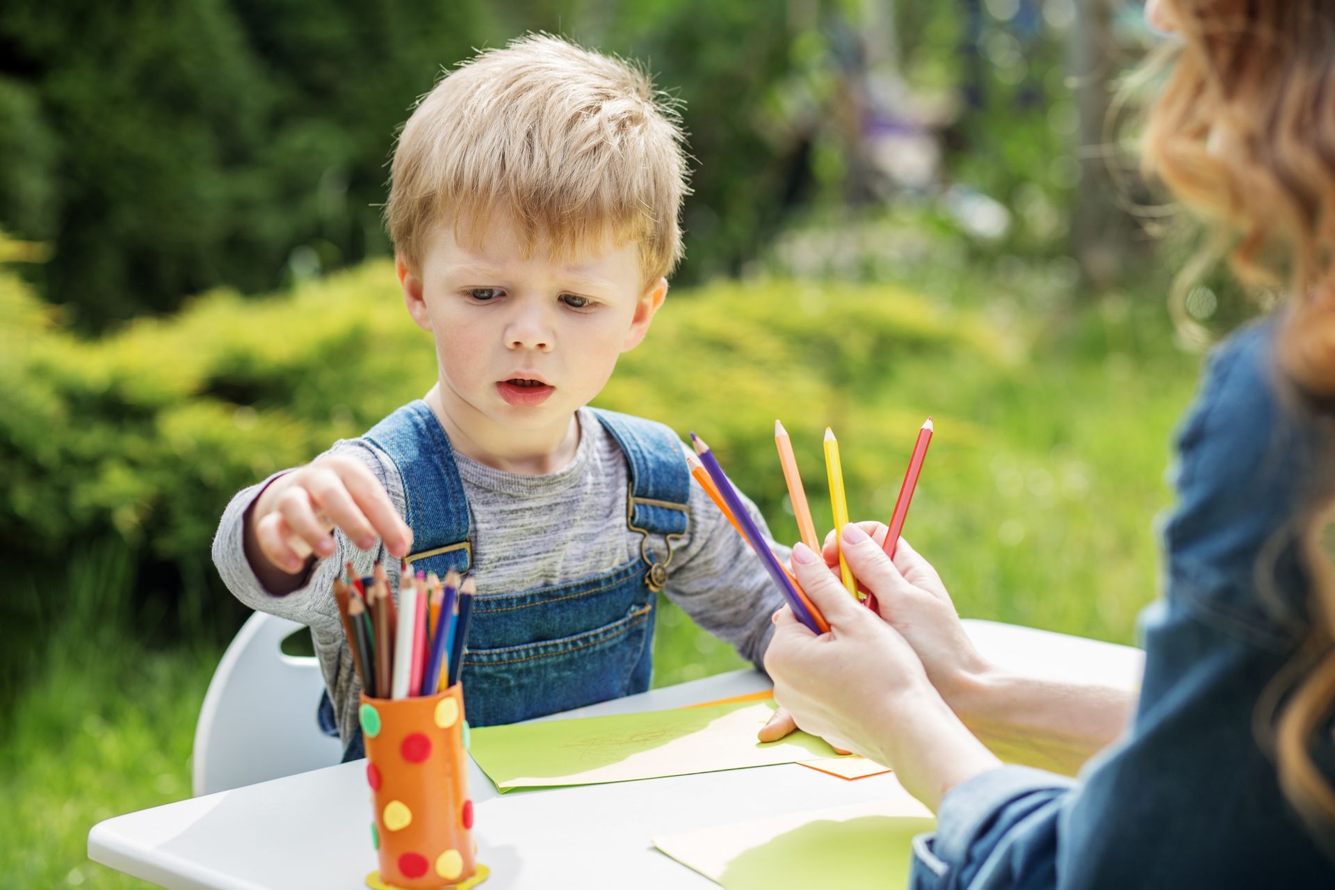 Boy seated outdoors, choosing colored pencils from woman's hand. White table with drawing paper.