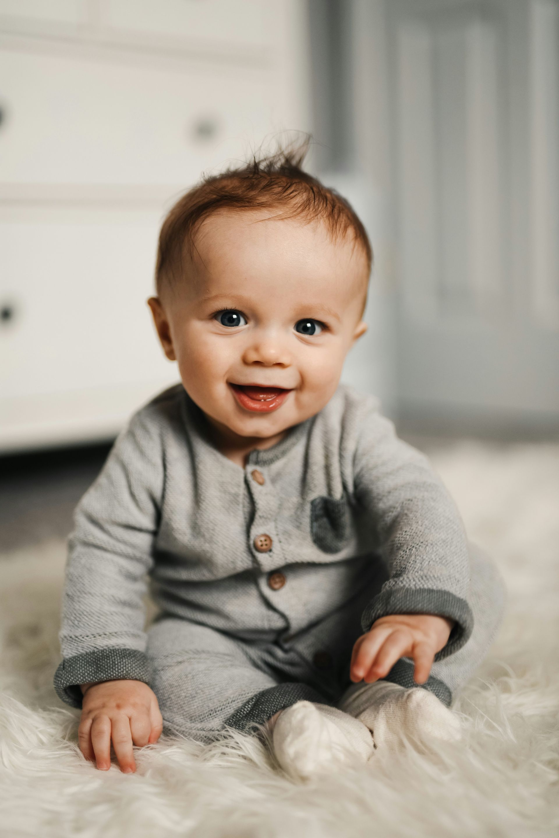 Smiling baby with spiky hair, wearing gray outfit, sitting on fluffy white rug.