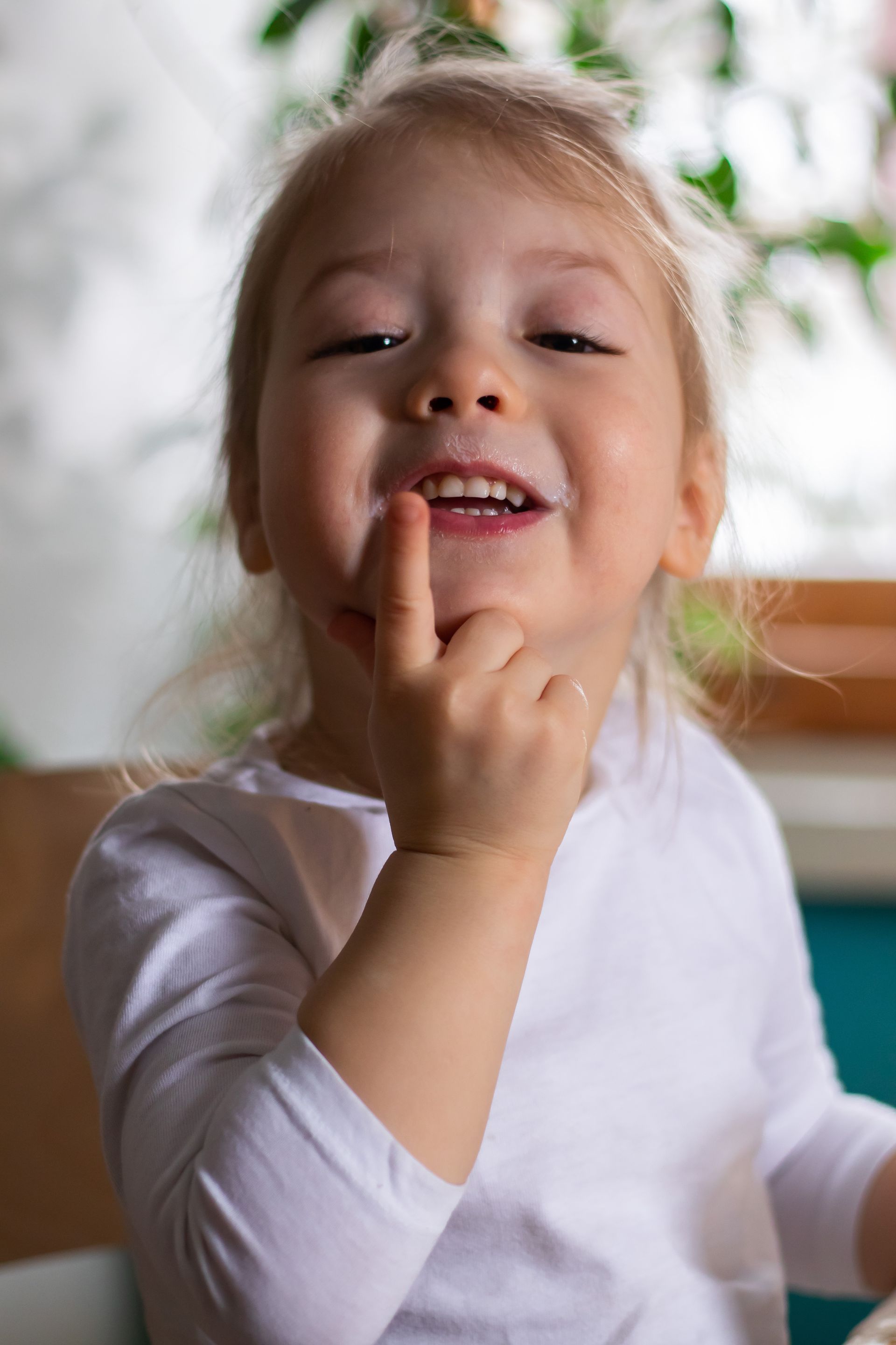 Smiling child with finger near mouth, wearing a white shirt.