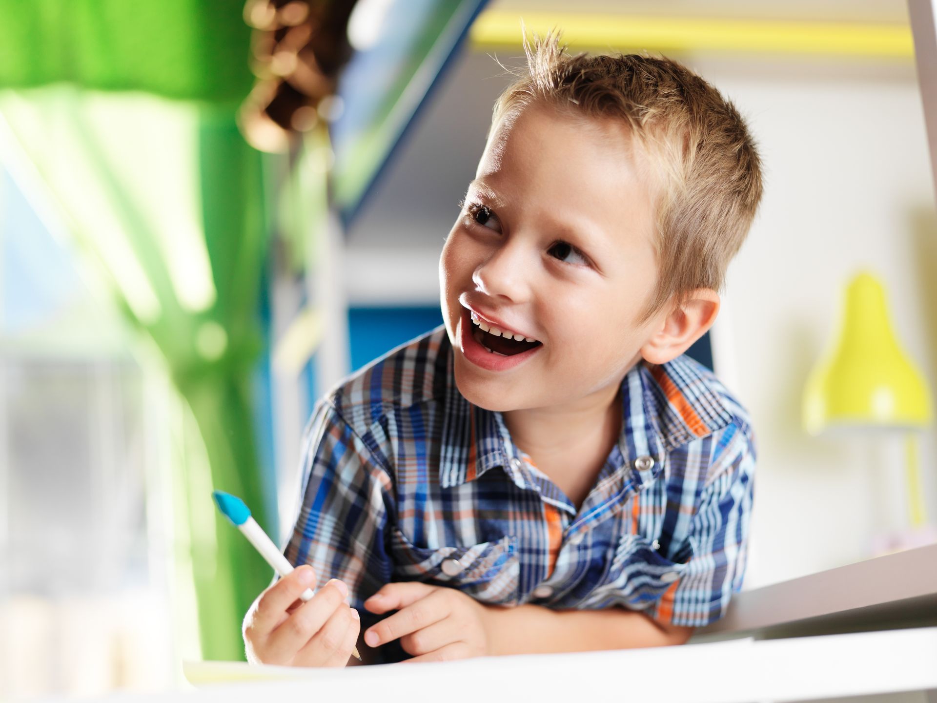 Boy with blond hair smiles widely, holding a blue marker, looking to the side.