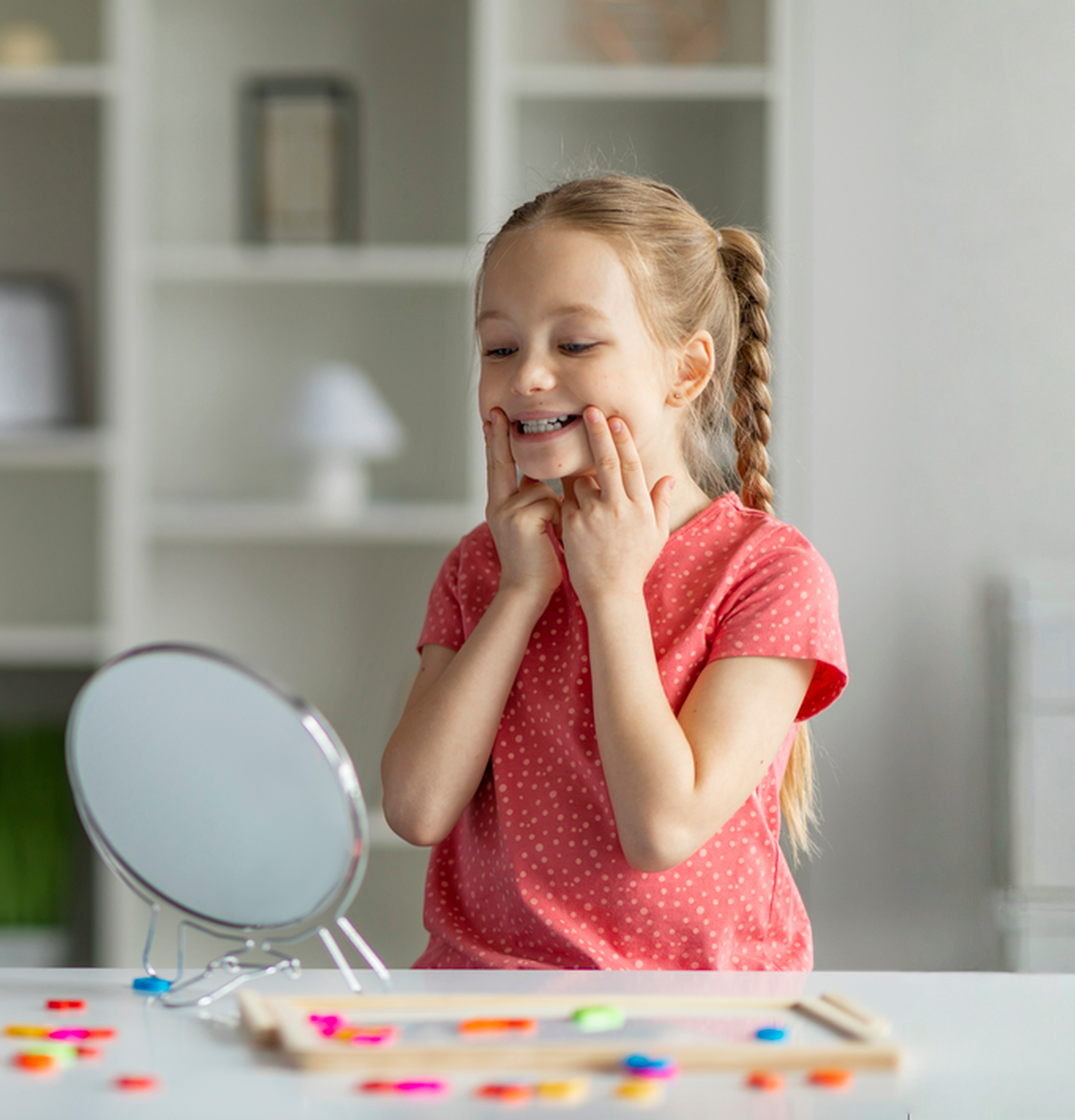 Girl smiles while looking in a mirror and touching her cheeks. A table has colorful shapes.