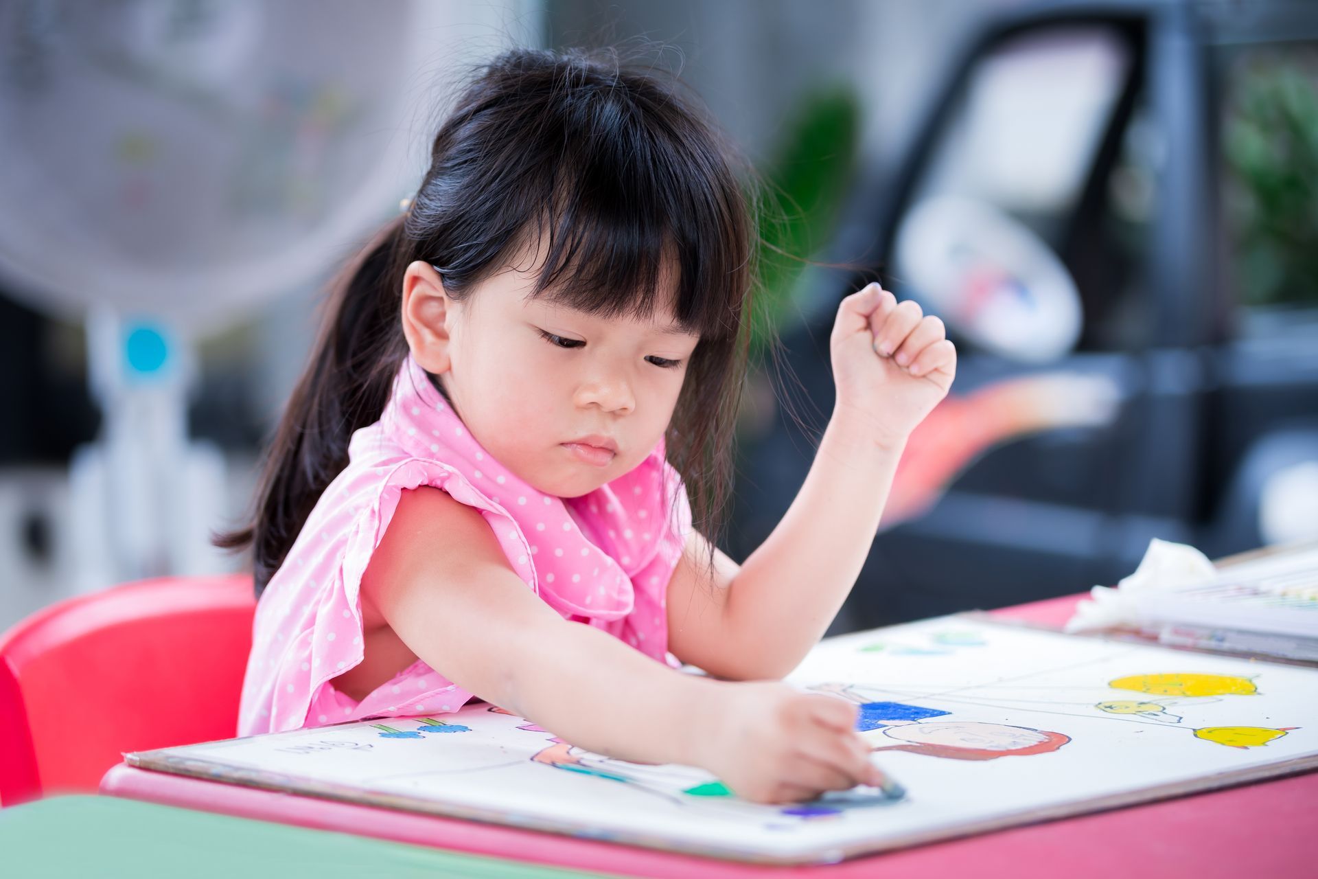 Girl in pink dress coloring at a desk, focused expression, indoors.