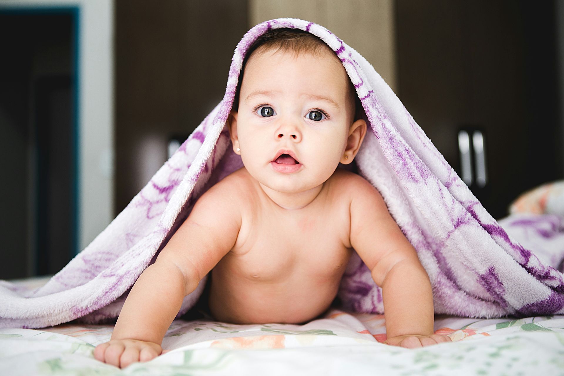 Baby under a purple floral blanket on a bed, looking surprised.