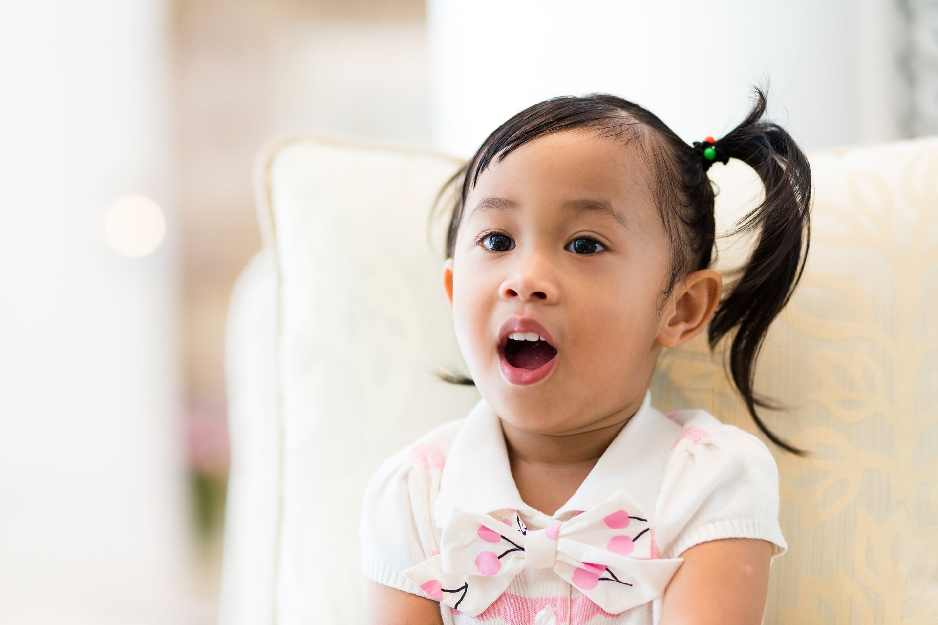 Young child with mouth open, sitting on a light-colored couch. She has one pigtail and a light-colored bow on her shirt.
