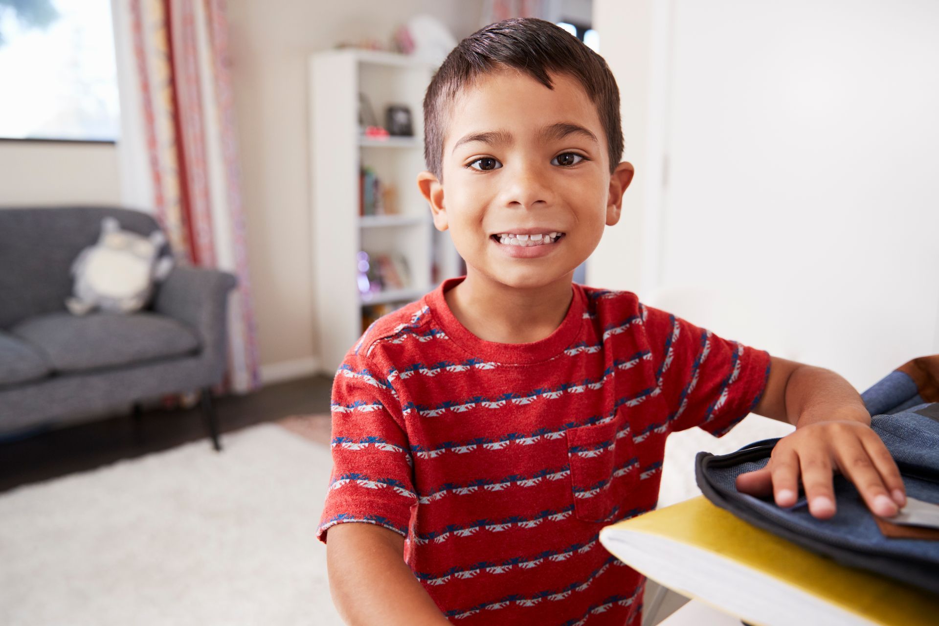 Smiling child at a desk with a backpack, wearing a red striped shirt.