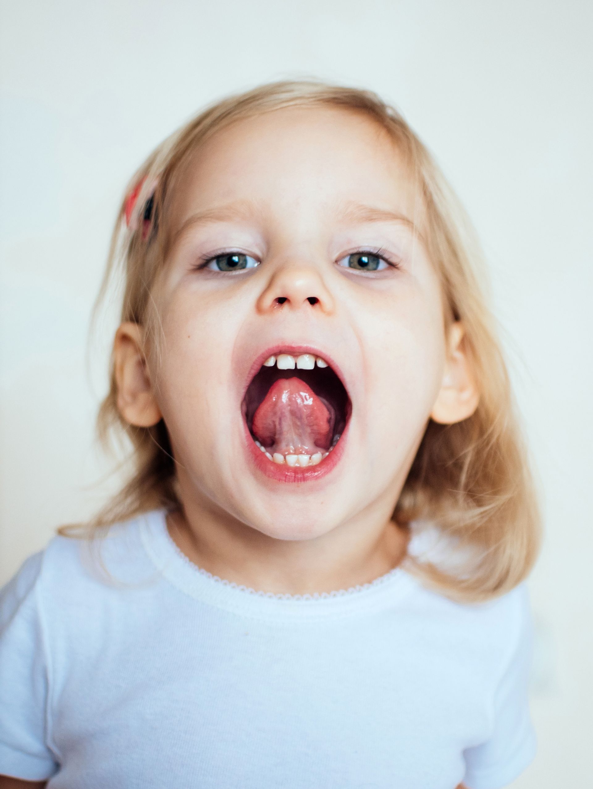 Blond toddler with mouth wide open, showing teeth and tongue; white shirt, light background.