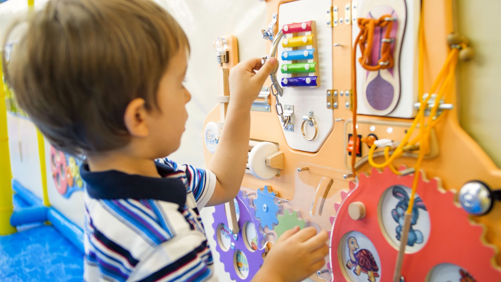 Boy playing with a wooden sensory board, exploring various latches, gears, and textures.