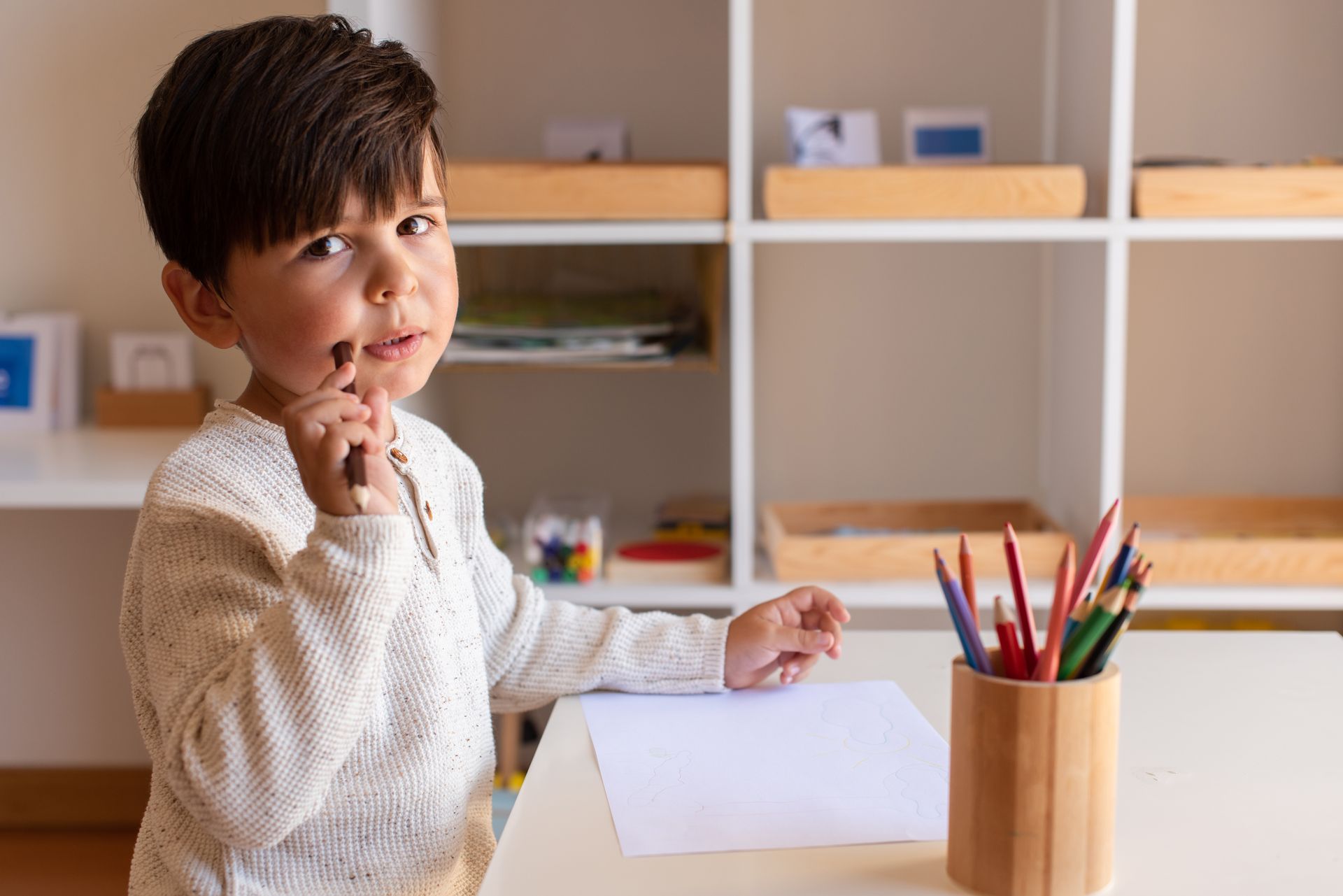 Boy at table, pencil to mouth, looking thoughtful. White paper and colored pencils on table in a classroom.