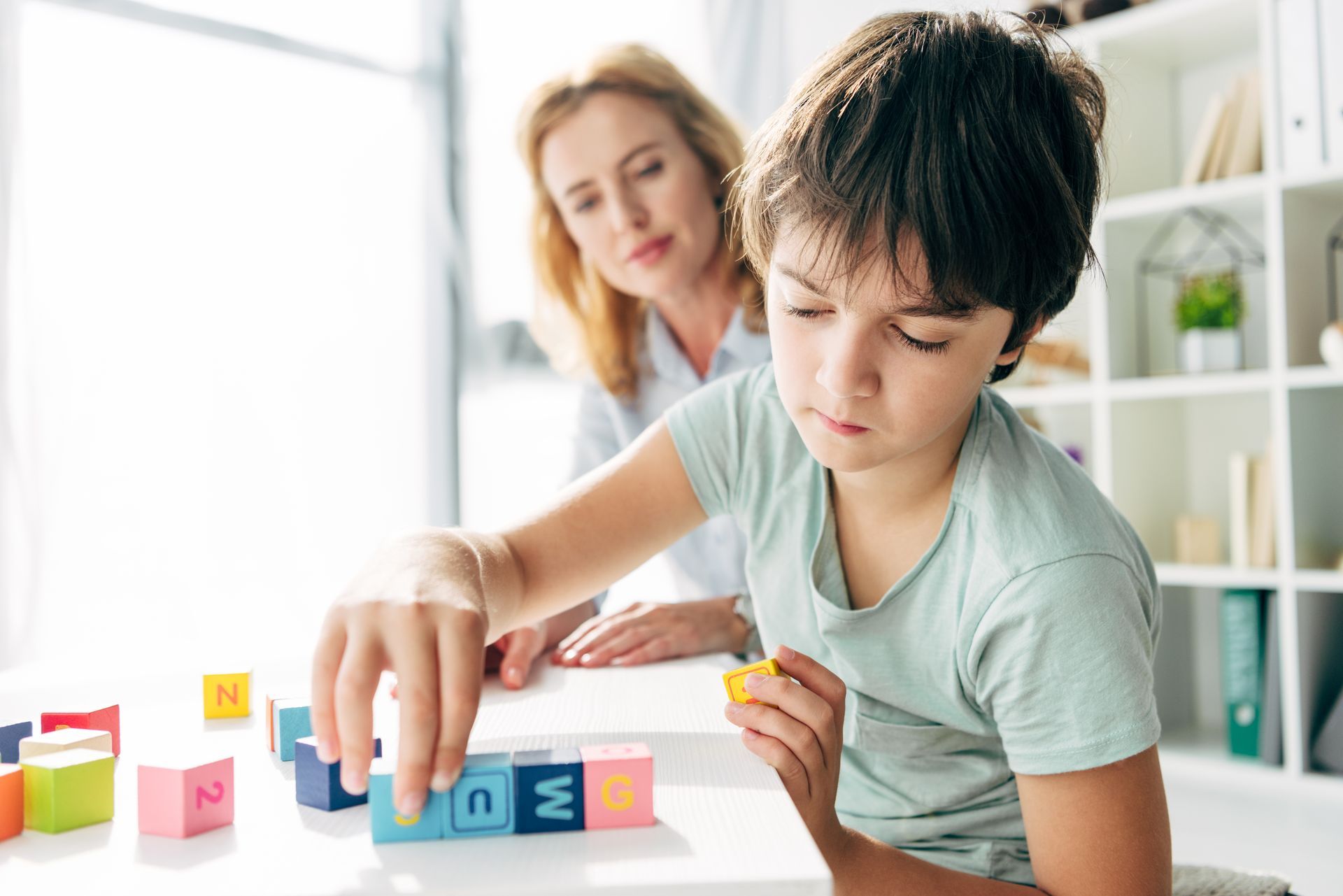 Boy focused on letter blocks, adult watches. Bright room, playing with blocks on a table.