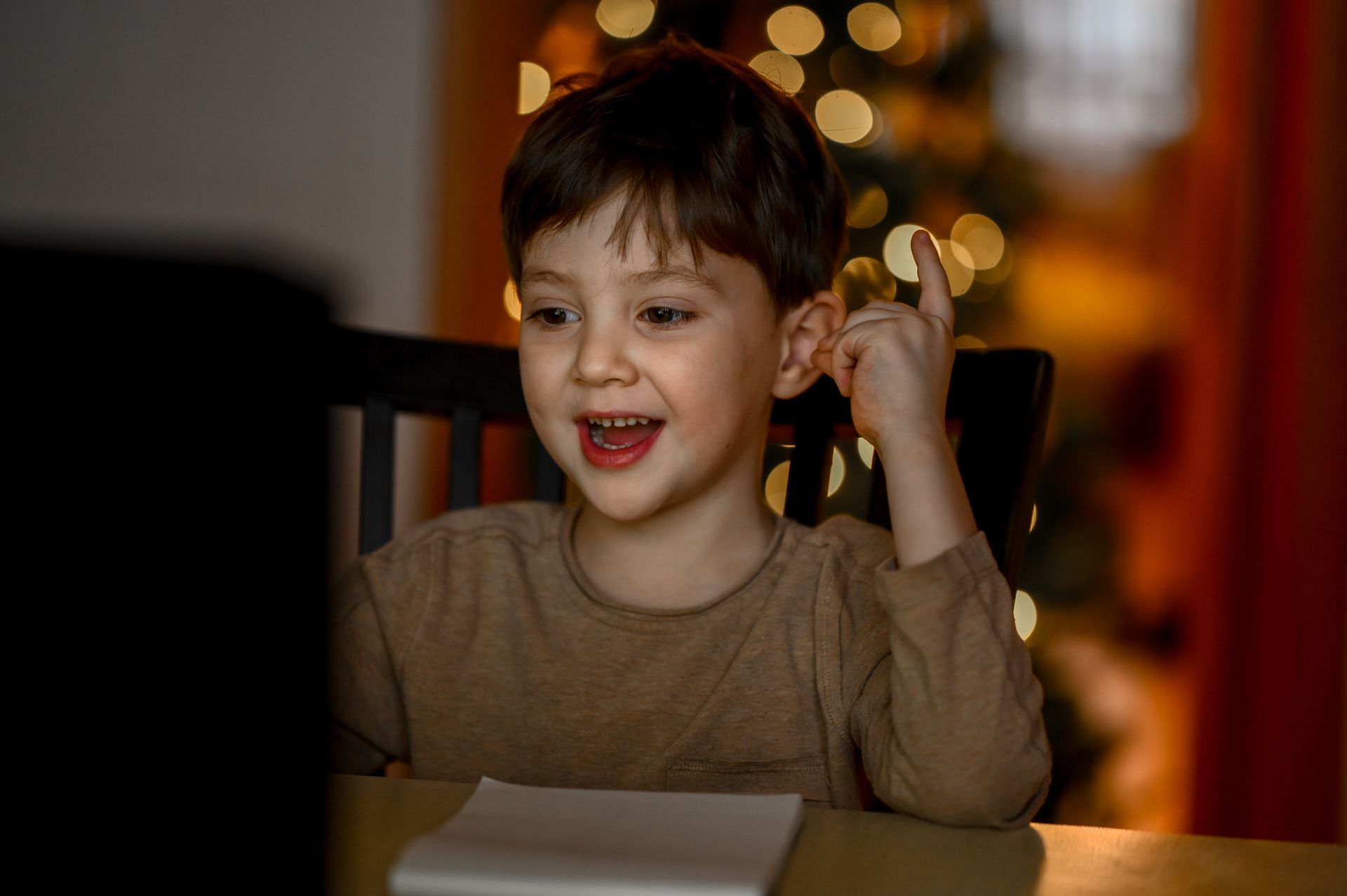 Young boy smiles with a raised finger, looking at a laptop with a Christmas tree background.