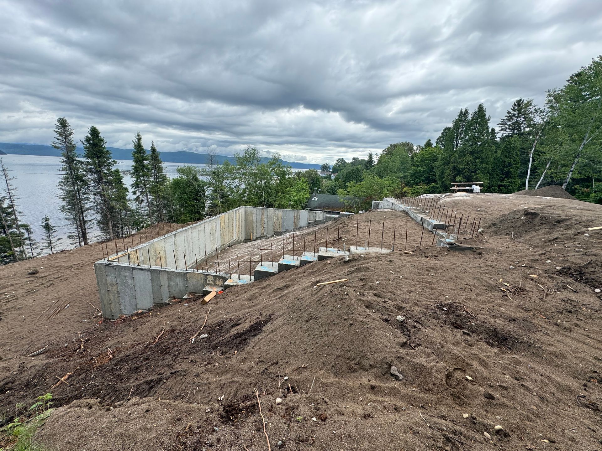 Un grand mur de béton est en cours de construction au sommet d'une colline de terre à côté d'un plan d'eau.
