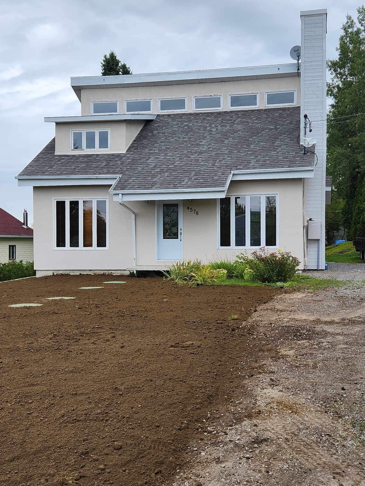 Une maison blanche avec un toit gris et beaucoup de terre devant.