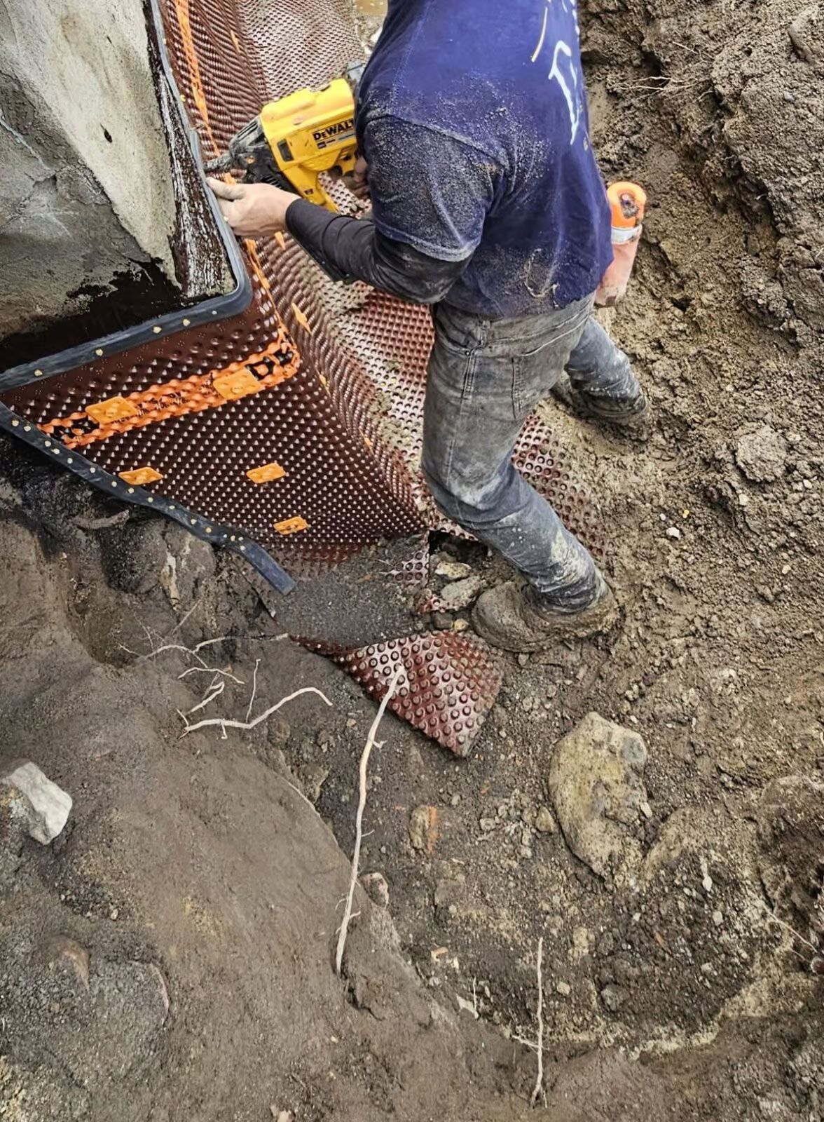 Un homme installe une membrane de drainage à picots sur un mur de fondation. Chantier extérieur. Tons bruns et beiges.
