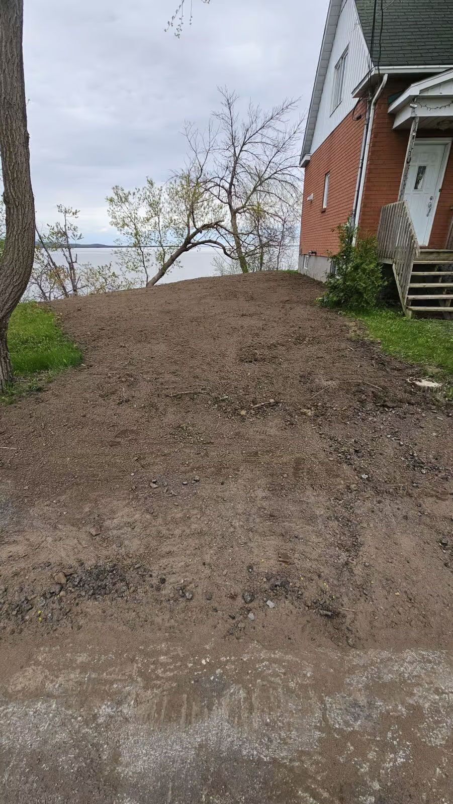 Tas de terre près d'une maison, probablement pour des travaux d'aménagement paysager. Vue sur un plan d'eau en arrière-plan.