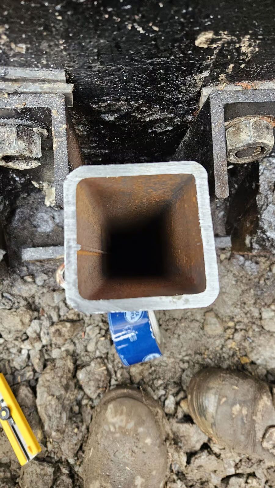 Vue de dessus d'un tube métallique carré enfoui dans la terre, probablement destiné à un chantier. Un niveau et une chaussure sont visibles.