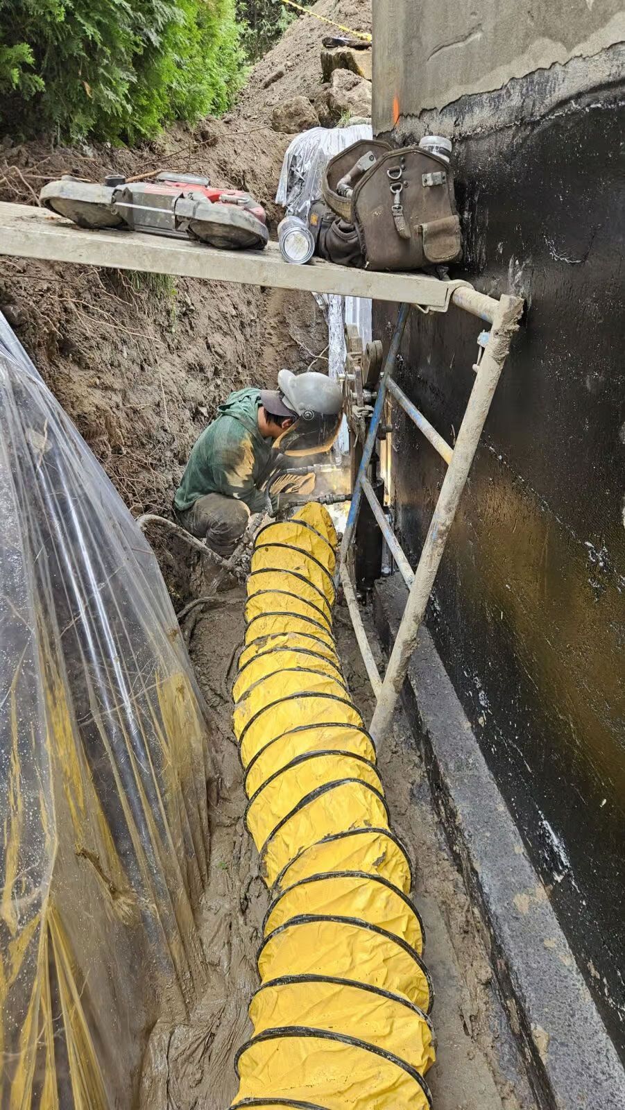 Un tuyau jaune longe une tranchée près d'un mur. Des matériaux et du matériel de construction sont présents.