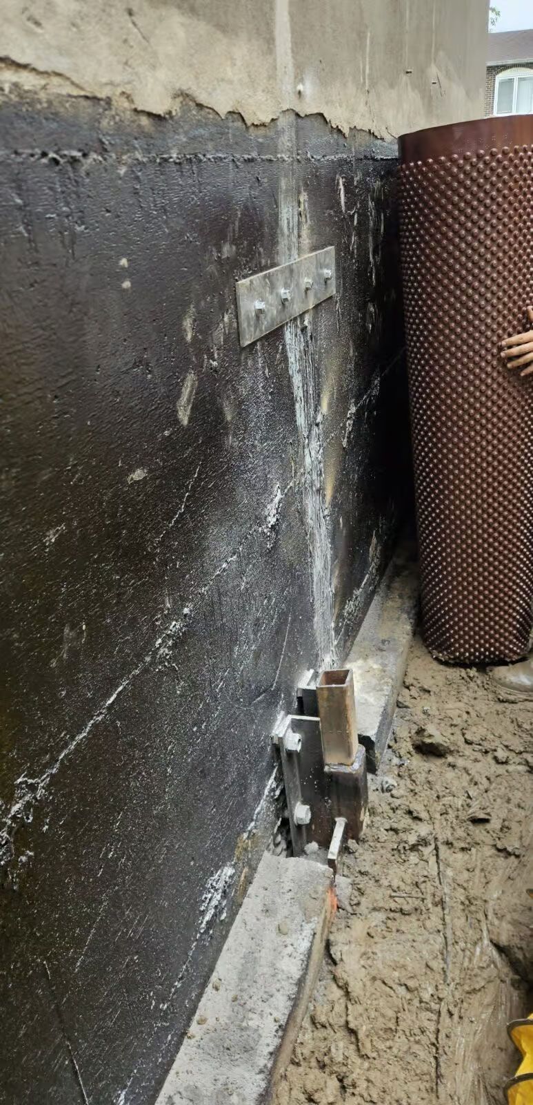 Membrane d'étanchéité noire fixée à un mur par des supports métalliques et un panneau de drainage à picots ; un chantier de construction.