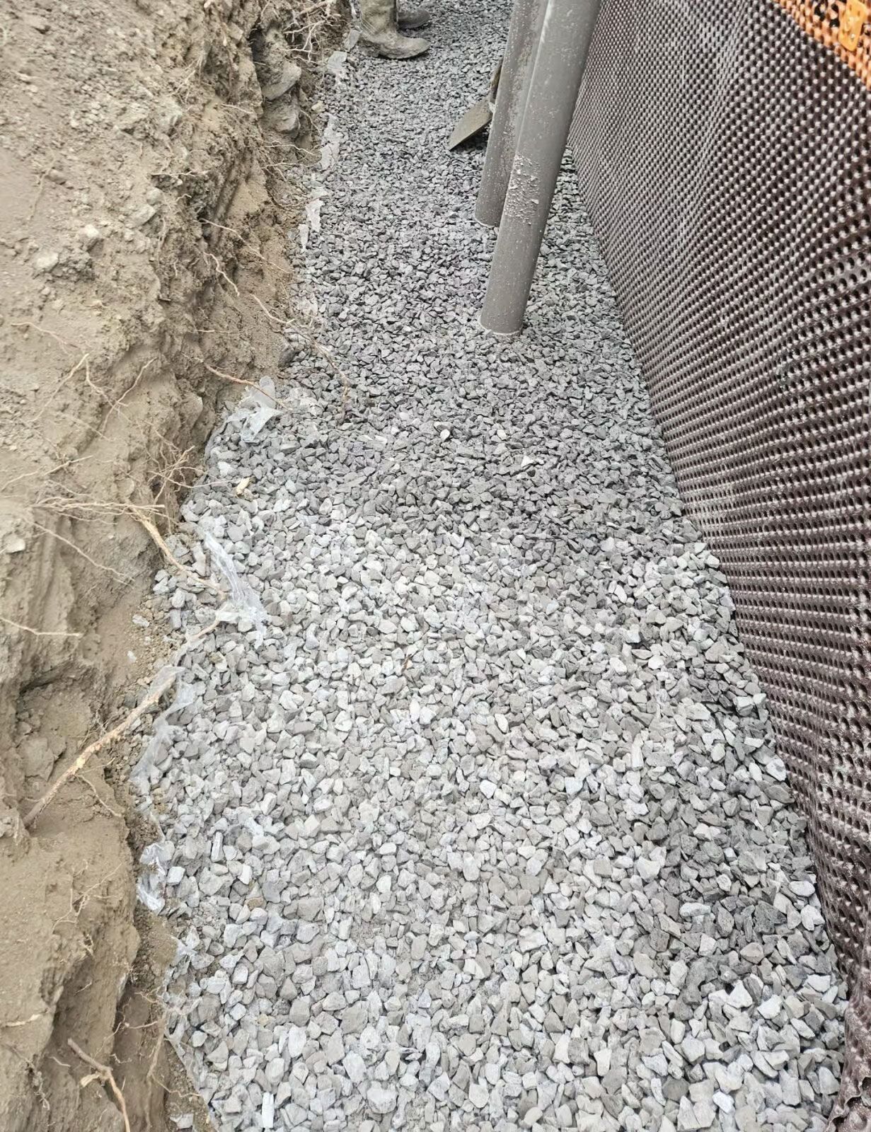 Tranchée remplie de gravier, jouxtant un mur de fondation en tôle ondulée noire et de la terre. Une canalisation est également visible.