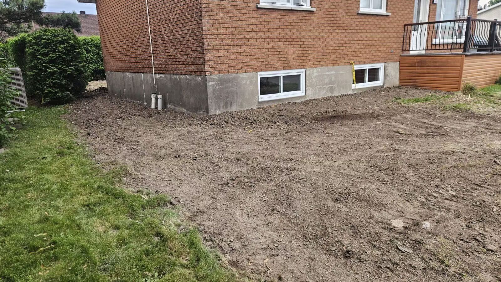 Une maison en briques aux fondations apparentes et à la terre nue devant. De l'herbe verte sur le côté.