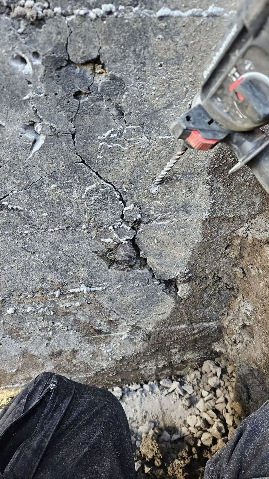 On utilise une perceuse sur du béton fissuré. La perceuse est grise et rouge. On aperçoit la lumière du soleil.