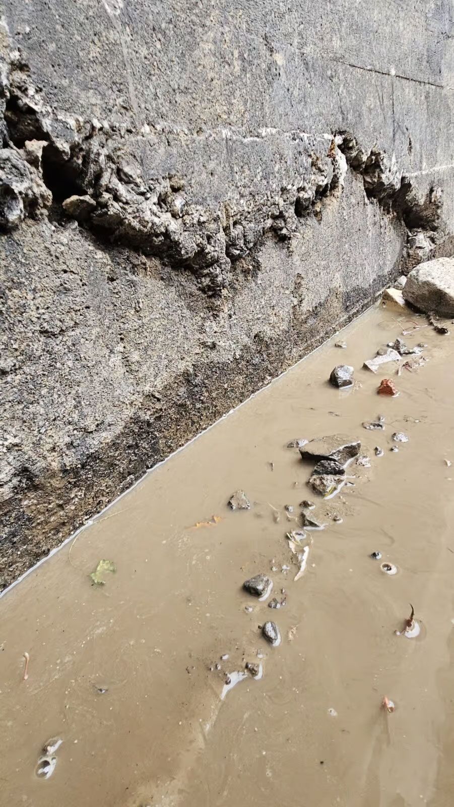 Mur en béton endommagé à côté d'eau boueuse avec des débris.