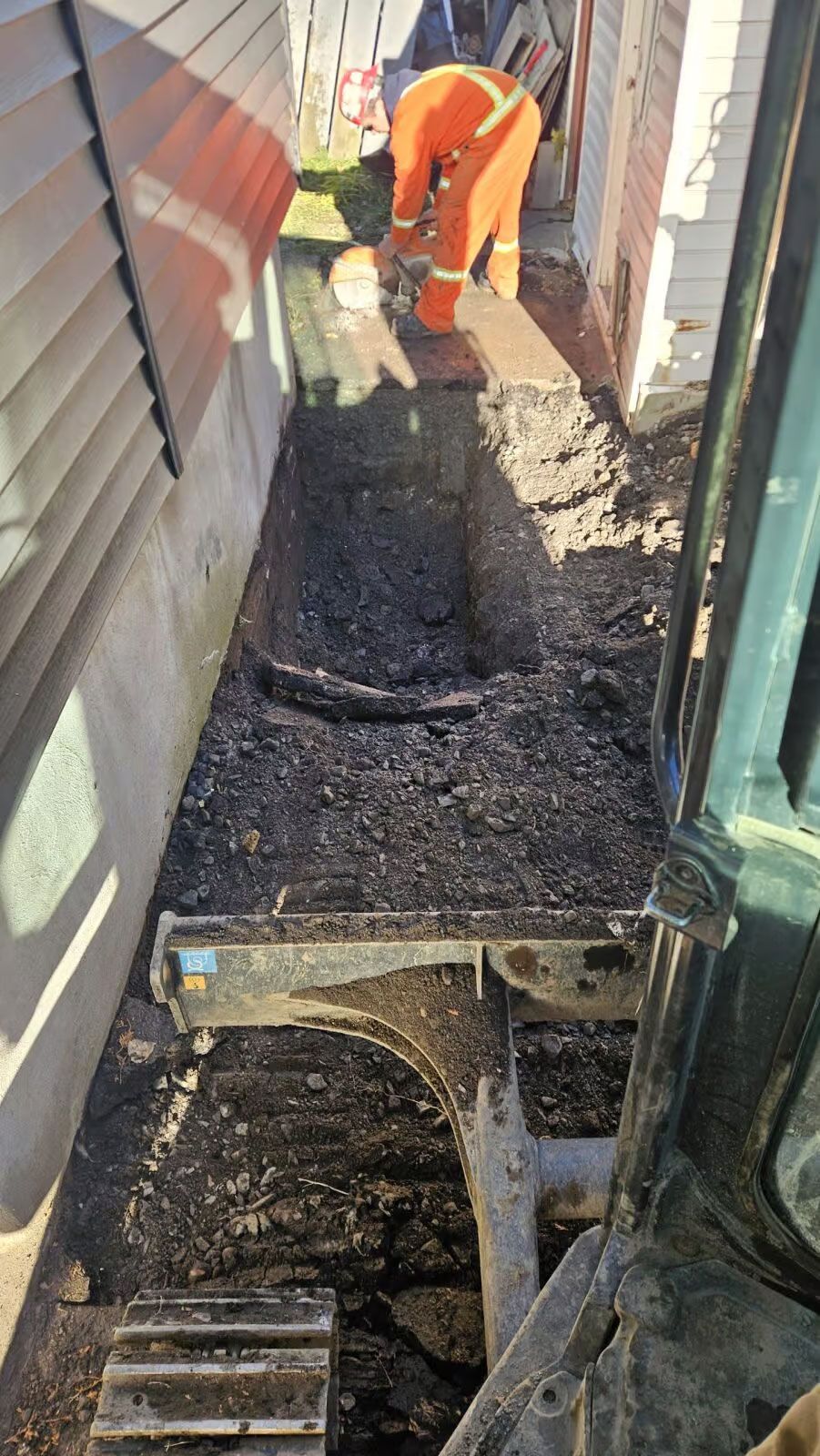 Une pelleteuse creuse une tranchée le long d'un bâtiment. Une personne en salopette orange se tient dans la tranchée.
