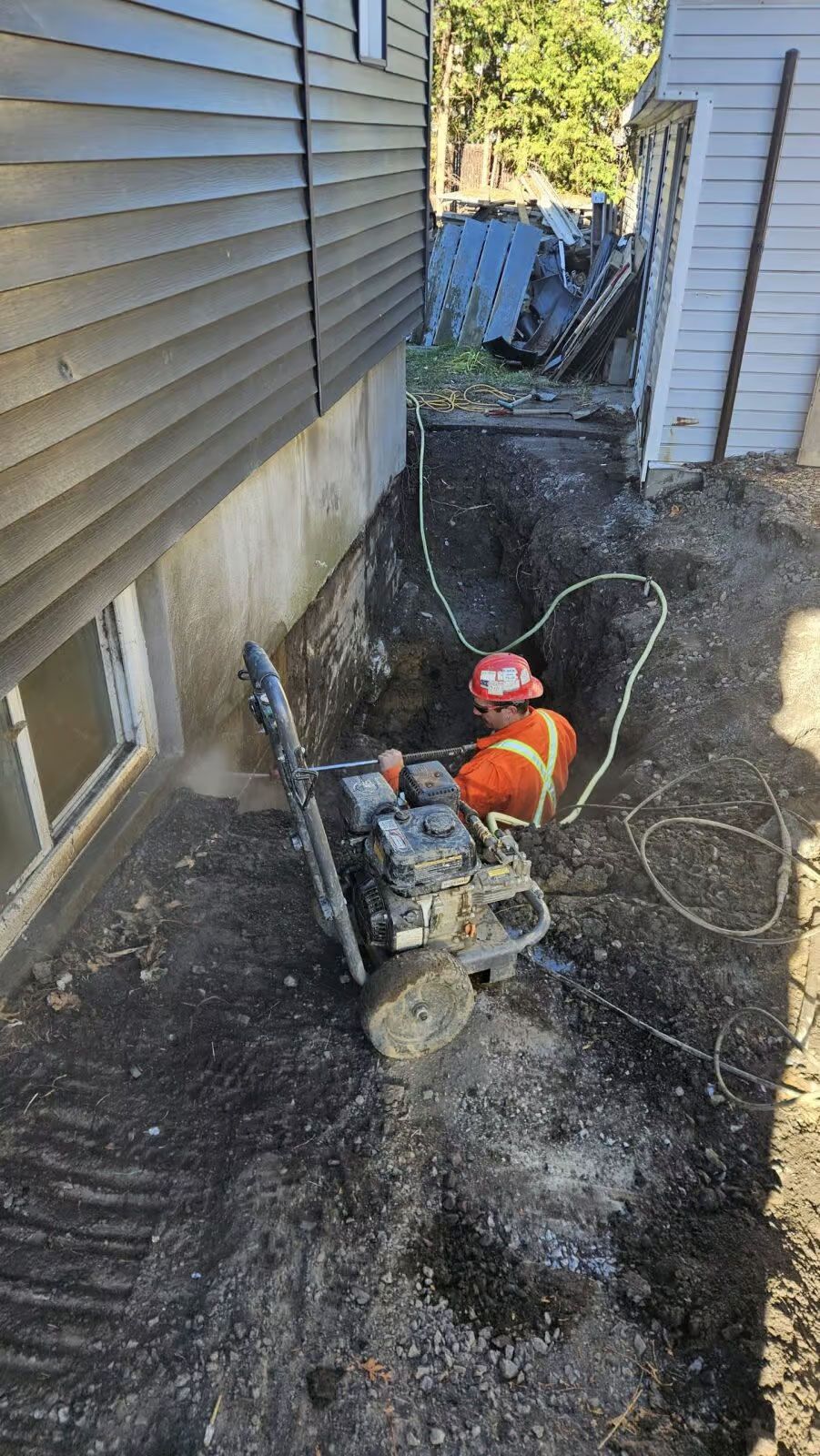 Une personne vêtue d'orange travaille avec une scie dans une tranchée à côté d'un bâtiment.