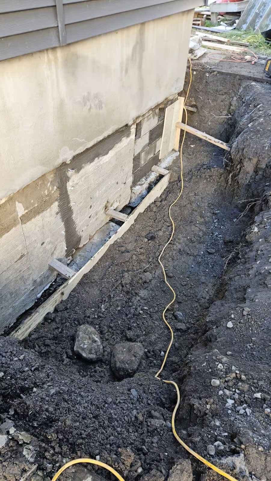 Mur de fondation excavé ; blocs de béton gris avec supports en bois et câble électrique jaune dans une tranchée de terre.