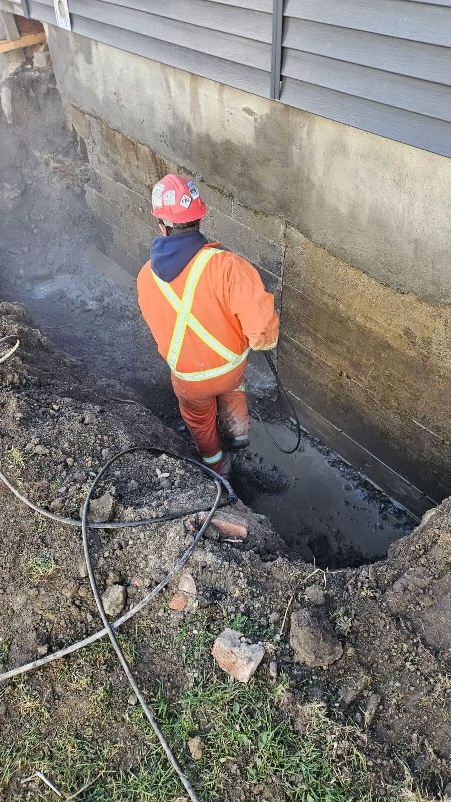 Ouvrier du bâtiment en combinaison orange et casque de chantier, travaillant dans une tranchée boueuse à côté d'un bâtiment.