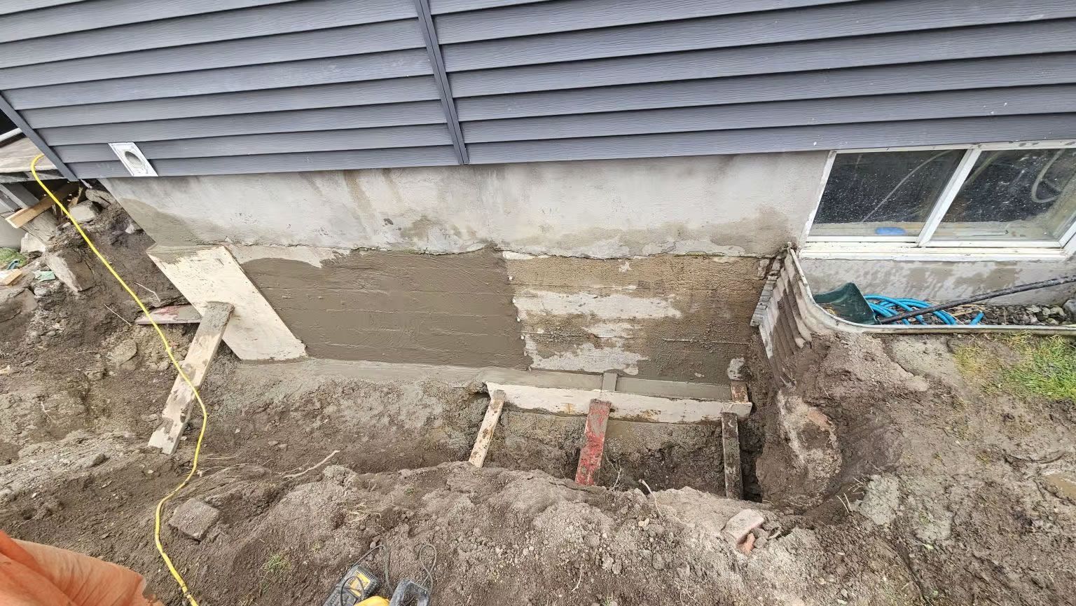 Travaux de réparation des fondations en cours ; terre, béton, supports en bois et revêtement extérieur visibles.