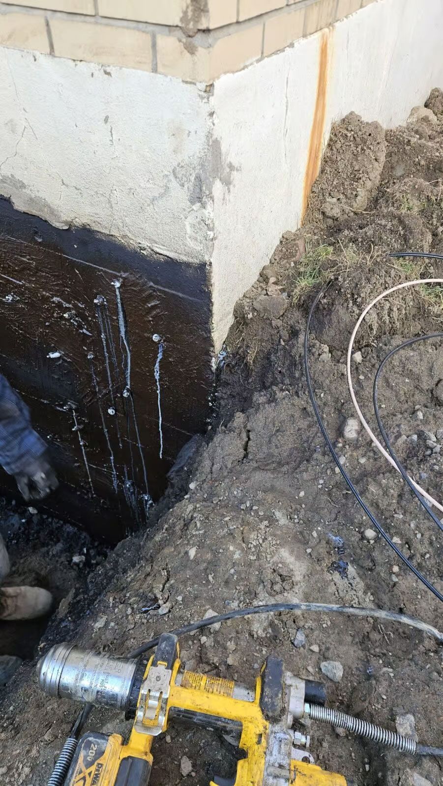 Réparation d'un coin de fondation ; enduit d'asphalte foncé sur béton, terre et outils jaunes visibles.