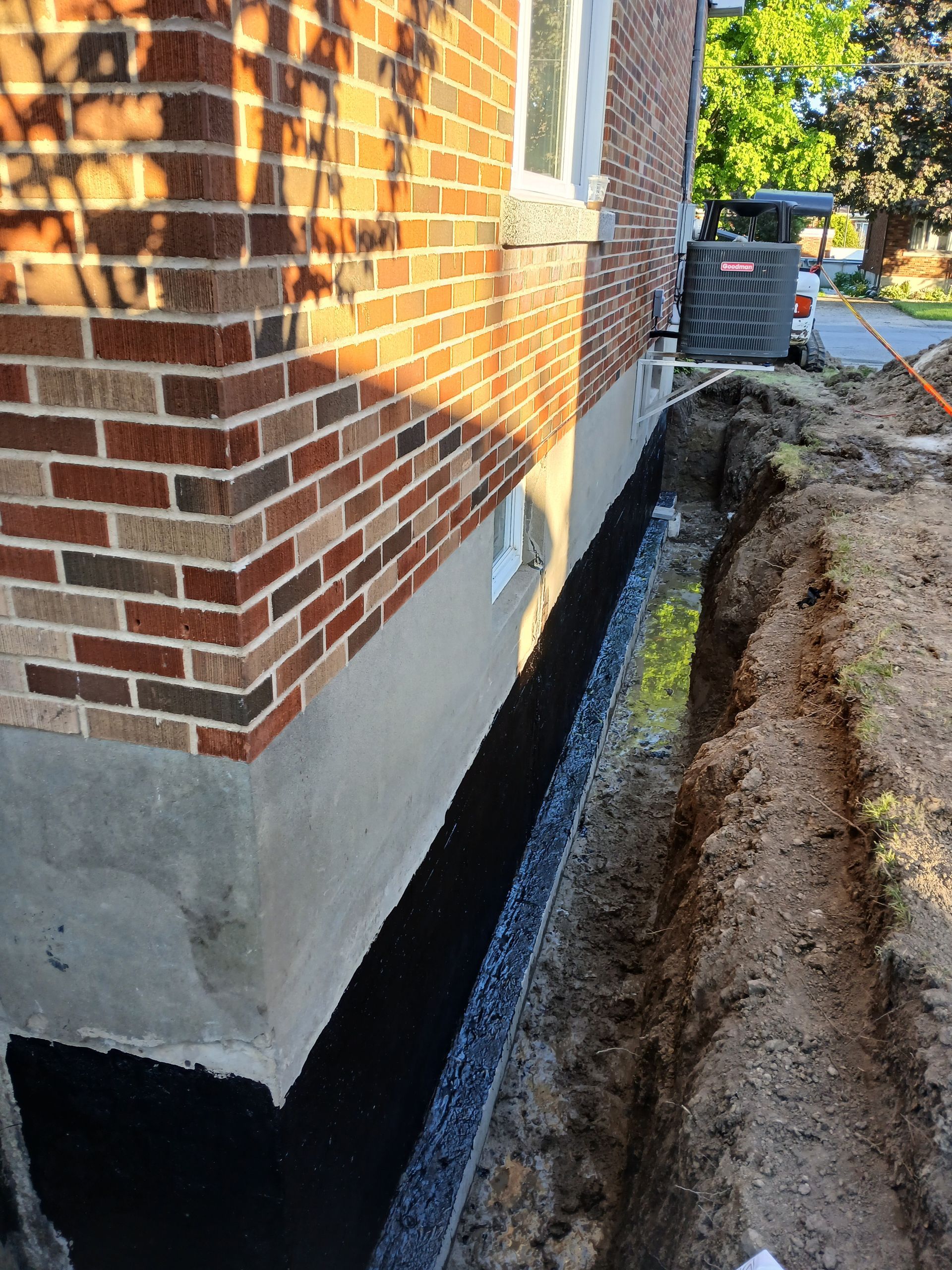 Vue extérieure des fondations d'une maison en briques avec une tranchée creusée à côté, probablement pour l'étanchéité.