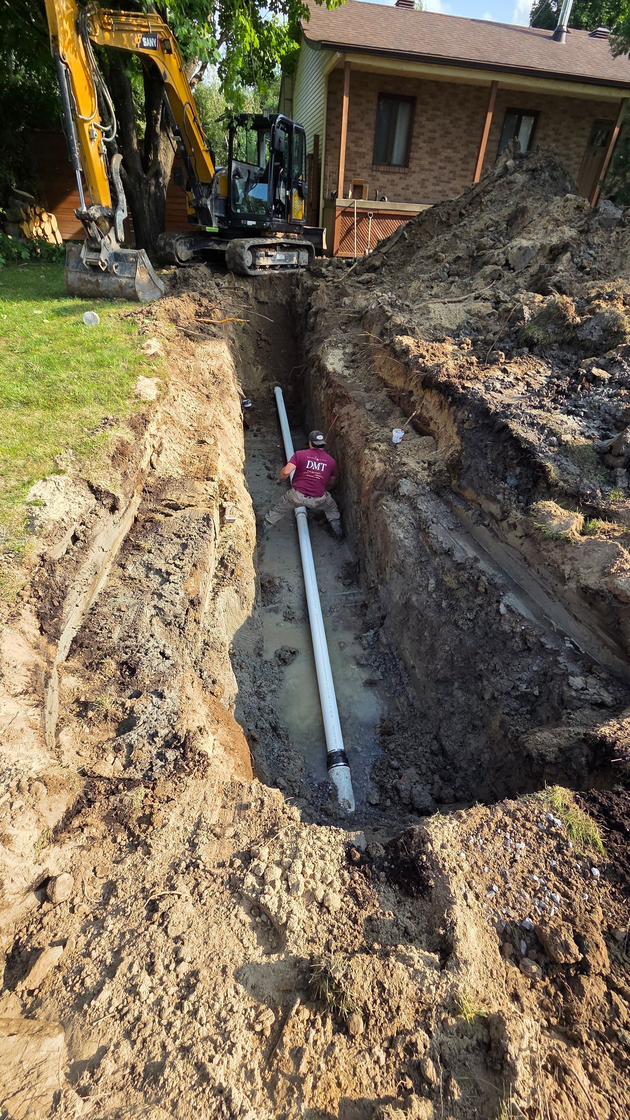 Un ouvrier dans une tranchée avec un tuyau blanc ; une excavatrice et un bâtiment en briques sont visibles.