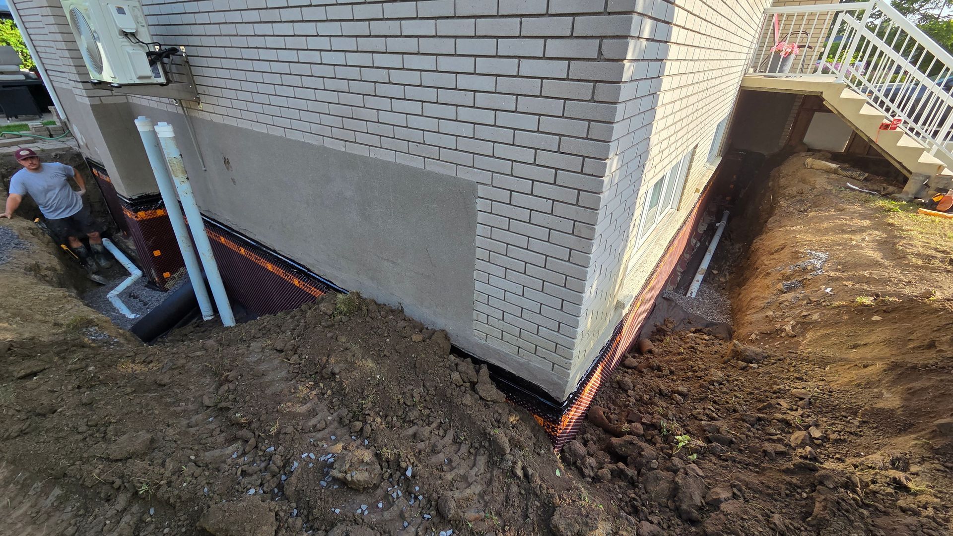 Chantier de construction avec travaux d'étanchéité des fondations. Bâtiment en briques beiges, tranchée et une personne qui observe.