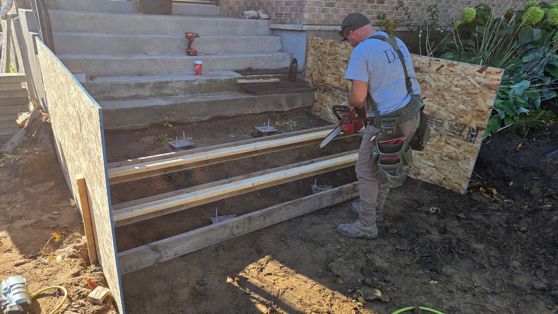 Une personne utilise une scie pour travailler sur un escalier extérieur en construction, composé de marches en béton et de bois de construction.