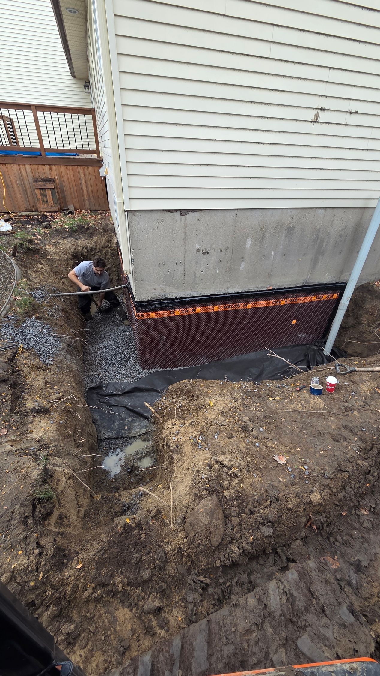 Excavation en cours du mur extérieur du sous-sol. Un ouvrier se trouve près des fondations apparentes. Terre, bâche noire et matériau de drainage orange.