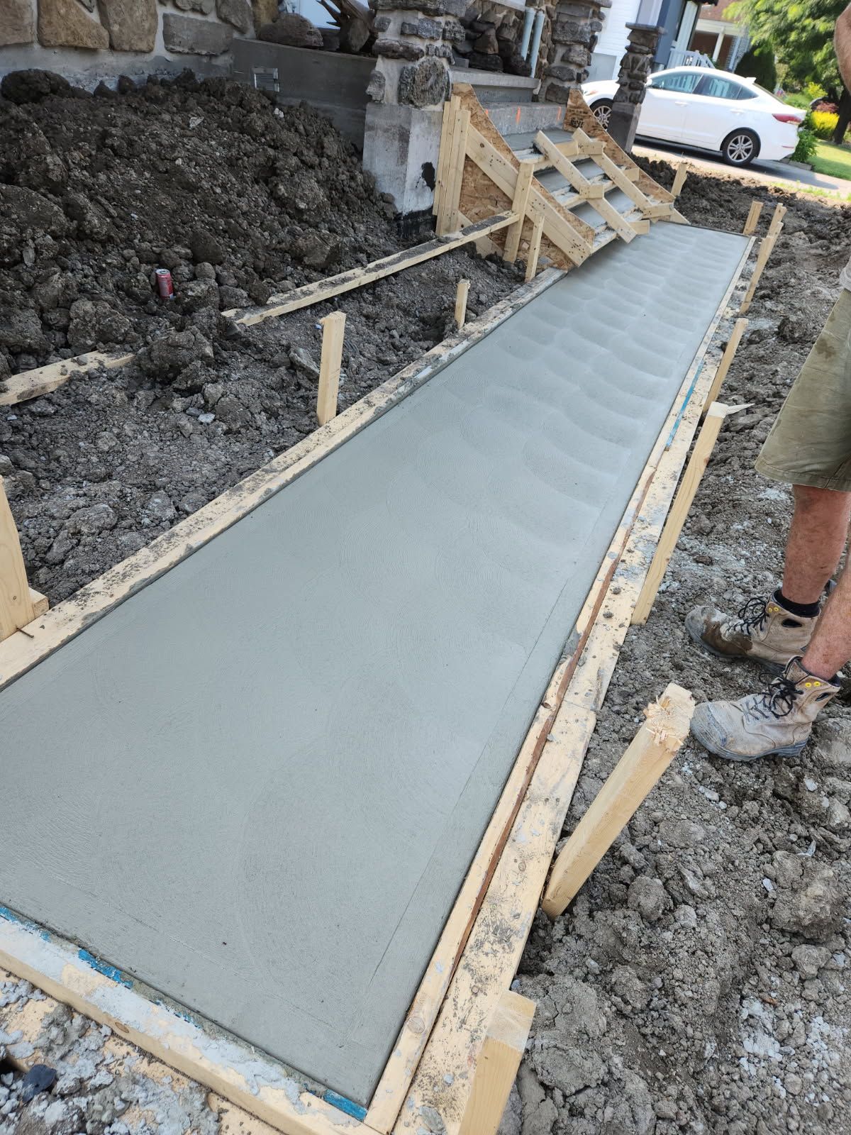 Trottoir en béton fraîchement coulé à l'intérieur de coffrages en bois. Le chantier est entouré de terre.