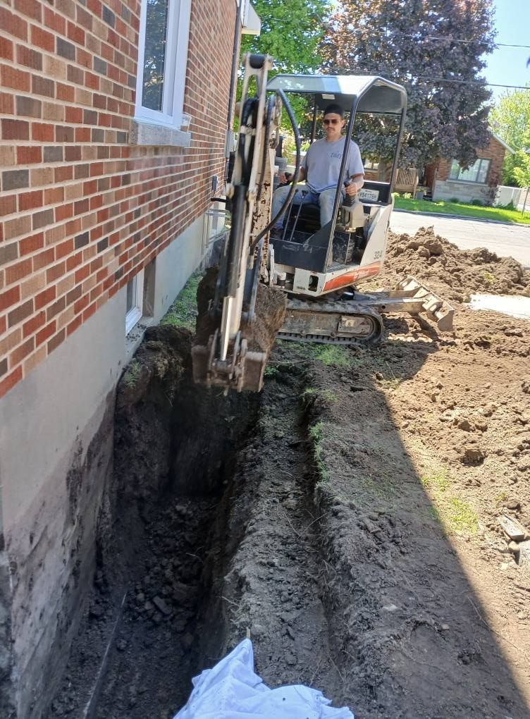 Une personne manœuvre une petite excavatrice en train de creuser une tranchée le long d'un bâtiment en briques.
