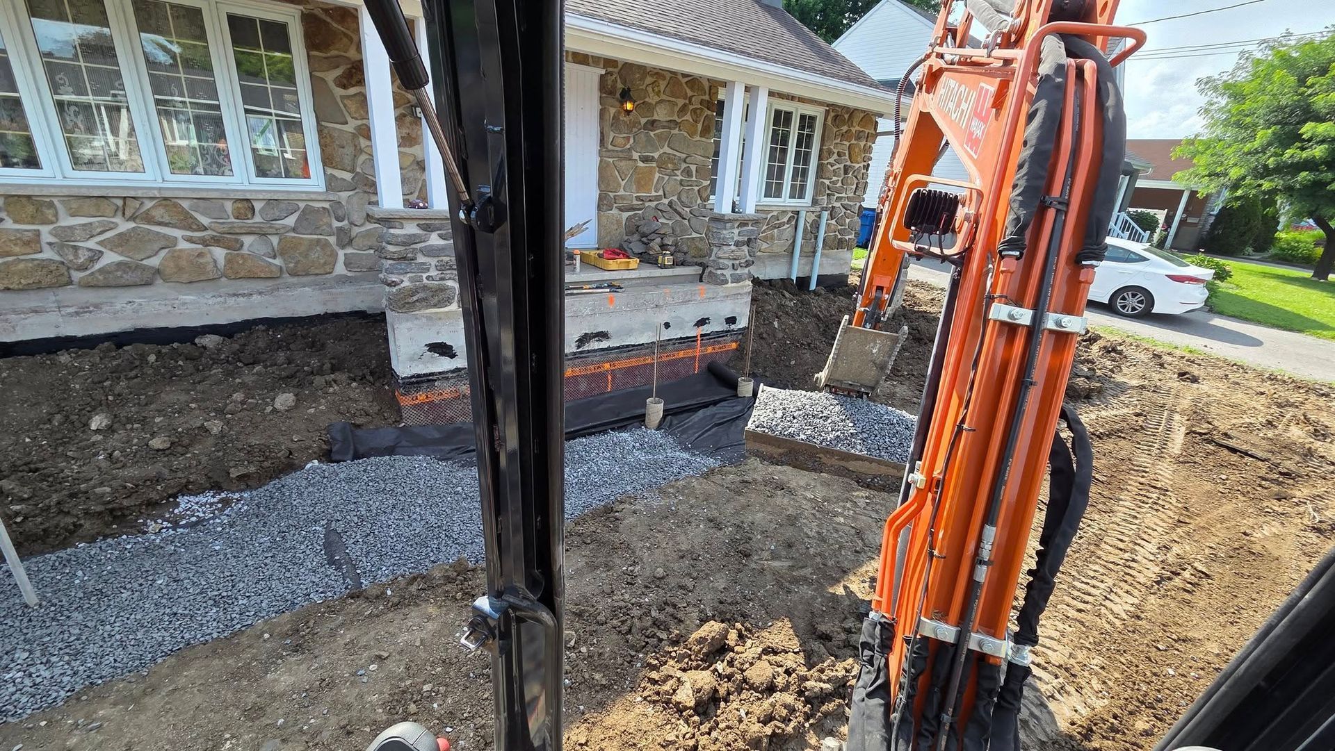Une excavatrice creuse près des fondations d'une maison, laissant apparaître du gravier et de la terre.